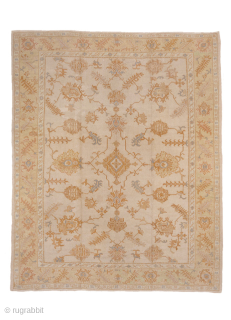 This antique Oushak rug offers timeless elegance with its soft ivory ground and warm golden motifs. The field is decorated with an allover pattern of stylized palmettes, vines, and geometric medallions, framed  ...