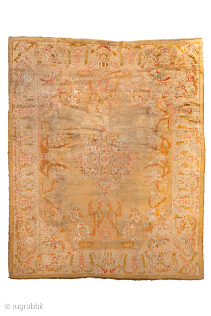 This antique Oushak rug features a warm, time-softened palette of gold, apricot, and blush tones, anchored by a central medallion design with playful floral and vine motifs. The wide border with decorative  ...