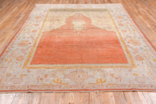 This antique Oushak rug showcases the beauty of Anatolian weaving with its striking open field in warm terracotta red, framed by soft blush, ivory, and golden tones. The spacious design and gentle  ...