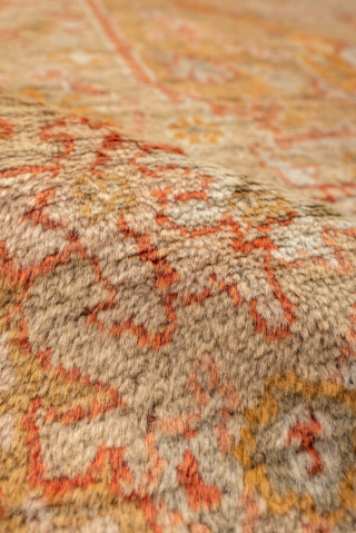 This antique Oushak rug features a warm, time-softened palette of gold, apricot, and blush tones, anchored by a central medallion design with playful floral and vine motifs. The wide border with decorative  ...