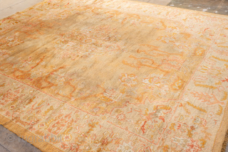 This antique Oushak rug features a warm, time-softened palette of gold, apricot, and blush tones, anchored by a central medallion design with playful floral and vine motifs. The wide border with decorative  ...