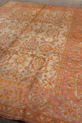 This antique Oushak rug, woven circa 1920, boasts a luminous palette of soft coral, apricot, and golden hues accented by subtle ivory highlights. Its allover pattern features stylized floral and geometric motifs  ...