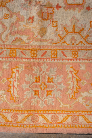 This antique Oushak rug, woven circa 1920, boasts a luminous palette of soft coral, apricot, and golden hues accented by subtle ivory highlights. Its allover pattern features stylized floral and geometric motifs  ...