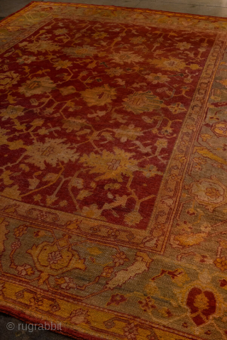 This antique Turkish Oushak rug, woven circa 1900, features a striking deep red field adorned with soft gold and muted blue floral motifs. The wide, decorative border frames the design beautifully, showcasing  ...